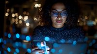 Young mixed-race woman with curly hair wearing glasses using smartphone at night with bokeh city lights creating digital glow effect on face and clothing.