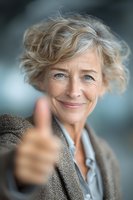 Confident mature businesswoman with short blonde hair giving thumbs up gesture, smiling at camera in professional setting for success and approval concepts.