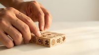 Hands arranging wooden cubes with engraved gear and puzzle icons on a light surface, symbolizing strategy, teamwork, and business solutions.