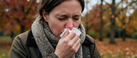 Middle-aged Caucasian woman blowing nose with tissue outdoors in autumn park. Seasonal allergies, cold, flu symptoms concept for healthcare advertising.