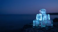 Stacked ice cubes with water droplets on dark rock surface against twilight ocean backdrop, suitable for beverage and refreshment marketing.