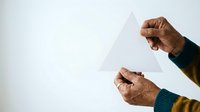 Close-up of male hands with medium-dark skin tone holding transparent triangle shape against white background for design, geometry, and educational concepts.