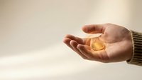 Hand holding glowing golden coin with warm light effect against soft beige background representing wealth, success, and financial opportunity.