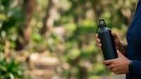 Black insulated water bottle held by person in blue jacket outdoors with blurred green foliage background, suitable for beverage, lifestyle and outdoor activity marketing.