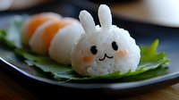 Cute bunny-shaped rice ball with carrot and nori details served on green leaf alongside sushi rolls on black plate, creative Asian cuisine presentation.