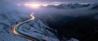 Winding mountain road with light trails at sunset, snow-covered peaks and misty valley below, aerial landscape photography for travel and nature projects.