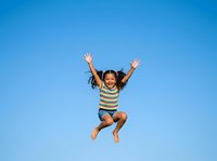 Joyful young girl with curly hair jumping with arms raised against clear blue sky, wearing striped shirt. Happiness and freedom.