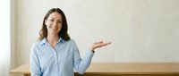 Professional businesswoman in light blue shirt presenting with open palm gesture in modern office space for corporate communication and marketing materials.