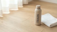 Spray bottle with train branding on wooden floor next to white towel and translucent containers, suitable for product mockups and wellness advertising.