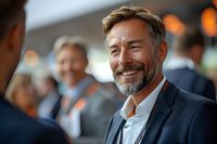 Confident middle-aged businessman with beard smiling during corporate networking event or business conference with colleagues in background.