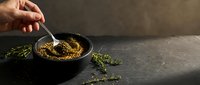 Hand stirring golden mustard sauce in black bowl with fresh herbs on dark stone surface for gourmet cooking and culinary preparation.