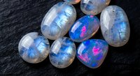 Polished rainbow moonstone gemstones with blue flash adularescence on dark slate surface for jewelry making and mineral collection displays.
