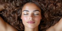 Young woman with closed eyes and serene expression lying down, surrounded by long curly brown hair spread out against dark background. Natural beauty concept.