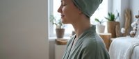 Young woman in green towel and robe relaxing after shower in bright modern bathroom with natural light and plants creating spa-like wellness atmosphere.
