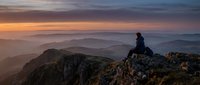 Silhouetted hiker sitting on rocky mountain peak at sunrise, overlooking misty valleys and distant hills in golden morning light for travel and adventure content.