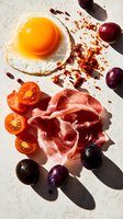 Sunny side up egg with prosciutto, cherry tomatoes, and fresh olives on white background. Bright morning light casting dramatic shadows. Food photography.