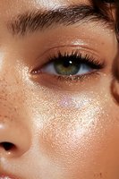 Close-up of glowing skin with shimmering makeup, green eye with long lashes and defined brow, featuring dewy complexion and freckles in golden light.