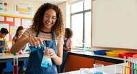 Young mixed-race female teacher conducting chemistry experiment in colorful classroom laboratory with students learning science education in background.
