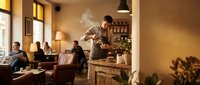 Professional male barista preparing coffee in cozy cafe interior with warm lighting and customers enjoying atmosphere in background.