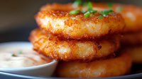 Crispy golden onion rings stacked with creamy dipping sauce on wooden surface, garnished with fresh herbs. Close-up view shows crunchy breaded coating.