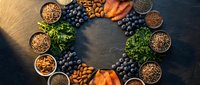 Superfood ingredients arranged in circular pattern with blueberries, nuts, seeds, leafy greens and colorful vegetables for healthy nutrition and wellness.