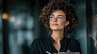 Professional young woman with curly hair and glasses smiling confidently in modern office environment for business and corporate presentations.