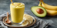 Creamy banana avocado smoothie in glass with fresh ingredients on rustic wooden plate against dark background, healthy breakfast option.