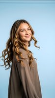 Young Caucasian woman with long wavy blonde hair smiling confidently in brown casual shirt against soft blue background for lifestyle and portrait photography.