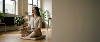 Young Asian woman practicing meditation in lotus position on yoga mat in bright modern living room with plants and natural light streaming through windows.