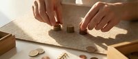 Hands of a young adult with medium skin tone arranging small wooden blocks and coins on a textured surface, demonstrating financial planning, budgeting, and money management concepts for educational or business use.