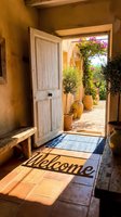 Rustic Mediterranean entryway with open white door, welcome mat, terracotta tiles, clay pots, blooming bougainvillea, and warm sunlight for home decor inspiration.
