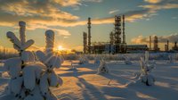 Industrial oil refinery plant silhouette at winter sunset with snow covered trees in foreground, dramatic orange sky and sunburst through factory towers.
