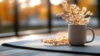 White ceramic mug with dried flowers on wooden surface against blurred window background, creating cozy autumn atmosphere with warm golden light.