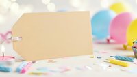 Blank kraft paper gift tag on colorful party table with balloons, confetti and celebration supplies for birthday events and festive occasions.