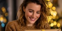 Young Caucasian woman with wavy hair smiling while looking down at glowing device, wearing cozy brown sweater against festive bokeh lights background.