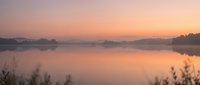 Serene lake at sunrise with misty morning atmosphere, calm water reflecting golden pink sky, forested shoreline, peaceful nature landscape for meditation and wellness projects.