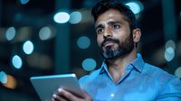 Young South Asian businessman using digital tablet at night, illuminated by blue bokeh lights in background, wearing casual blue shirt with trimmed beard.