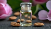 Essential almond oil in glass bottle with fresh almonds and pink flowers on dark wooden surface for natural skincare and aromatherapy treatments.