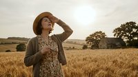 Woman in straw hat standing in golden wheat field at sunset, wearing floral dress and cardigan, looking upward with hand to hat brim, rural farmland landscape background.