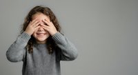 Young girl with curly brown hair covering eyes with hands in playful peek-a-boo gesture wearing gray knit sweater against neutral background.