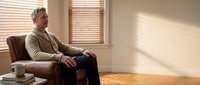 Middle-aged Caucasian man in beige sweater sitting relaxed in brown leather armchair by window with wooden blinds casting warm sunlight shadows across room.