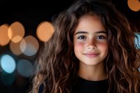 Young Hispanic girl with curly brown hair smiling warmly against dark background with soft bokeh lights creating magical atmosphere. Natural beauty portrait captures joy.