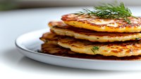 Stack of golden homemade pancakes garnished with fresh dill on white plate. Close-up view with selective focus shows fluffy texture and crispy edges.