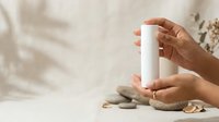 Hands holding blank white cylindrical product container with natural beige background, spa and wellness packaging mockup for cosmetics, skincare, or personal care branding.