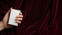 Unrecognizable person holding blank white box against dark burgundy velvet fabric background for product packaging mockup or branding presentation.