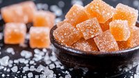 Orange fruit jelly cubes coated with sugar crystals in rustic ceramic bowl on dark background with scattered sugar, macro food photography.