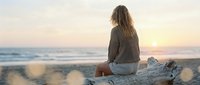 Young woman with long brown hair sitting on driftwood log watching golden sunset over ocean waves on peaceful beach during golden hour meditation moment.