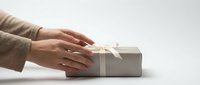 Female hands in beige sweater gently touching elegant gift box with white ribbon on clean white background for holiday celebrations and special occasions.