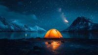 Illuminated orange tent on frozen lake shore reflects in ice surface against snowy mountain peaks under starry night sky with northern lights. Adventure camping scene.