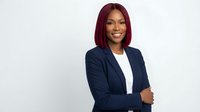 Professional African American businesswoman with burgundy bob haircut in navy blazer smiling confidently with crossed arms against white background.